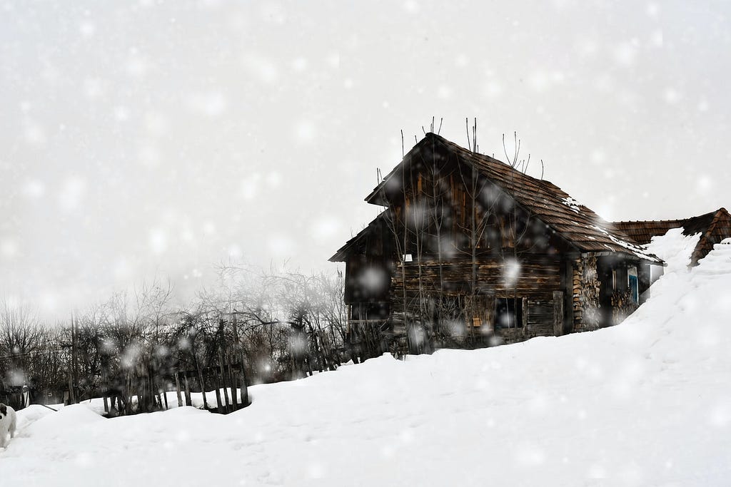 Photo of an old wooden cabin on a snowy hilltop beneath a grey cloudy sky.