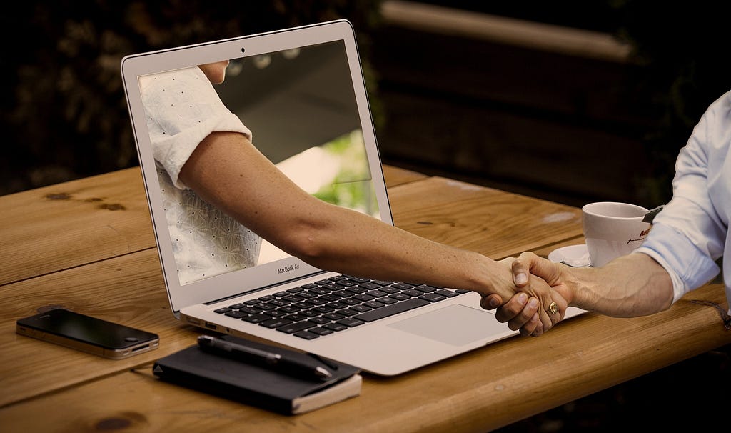 This image is meant to represent virtual collaboration. There’s a laptop on a desk. There’s a virtual arm coming out of the laptop screen shaking the real hand of the human using the laptop.