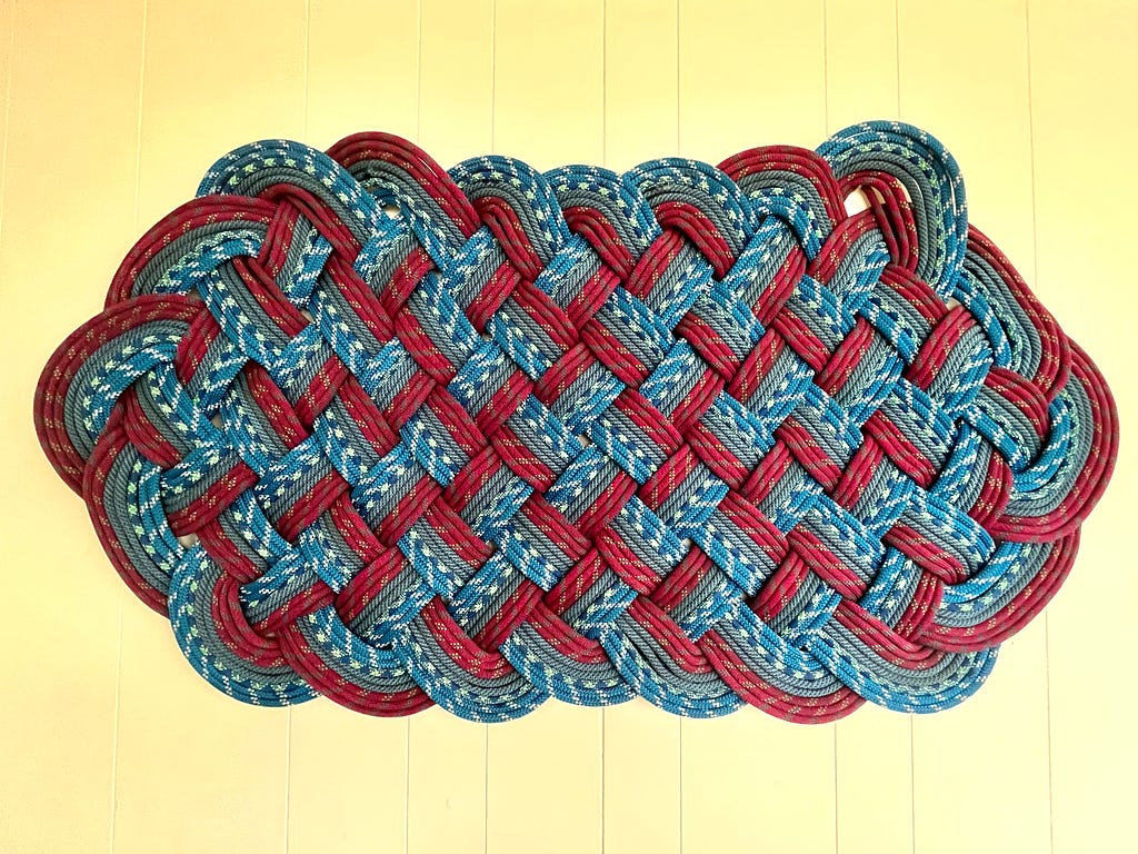 Color photo of a cream wooden wall background on which hangs a large ‘knotted’ sailing artwork in pale blue and dark red paracord rope, rectangularly wider than it is tall.