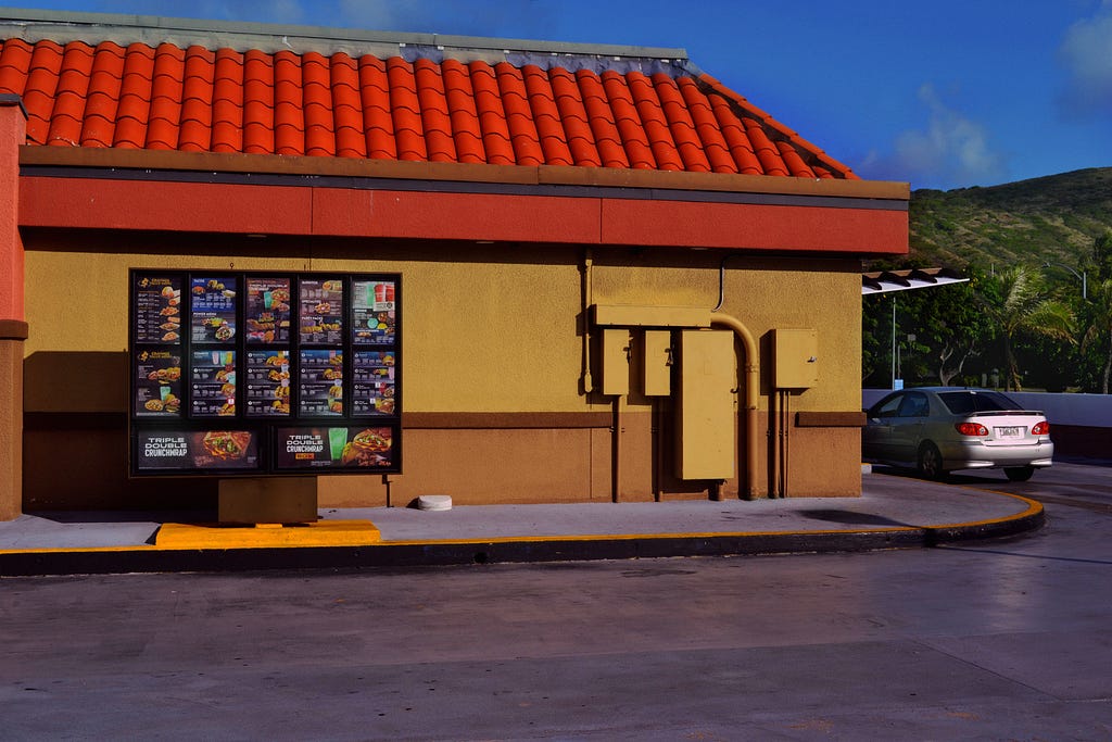 The side of a Taco Bell drive through in Hawaii Kai, Hawaii. Palm trees and a green mountain are in the background of the building and its drive thru menu.