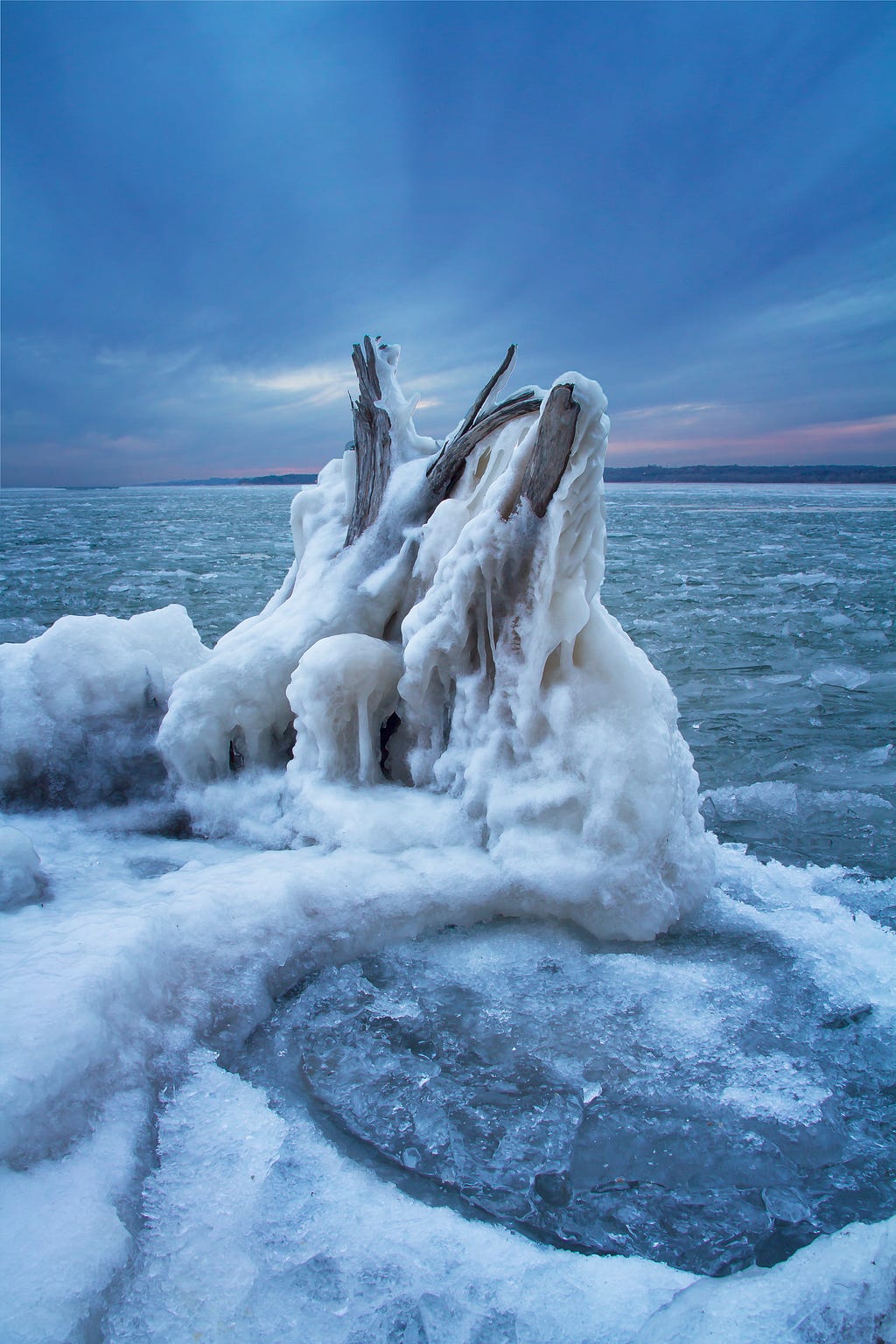 A stump of driftwood covered in ice and snow surrounded by a frozen lake with blue clouds and sunset.