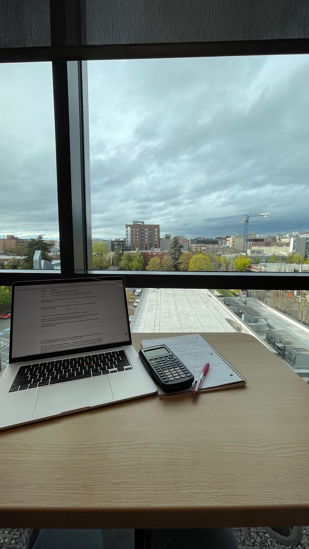 A laptop, notebook, and calculator on a wooden desk with large windows overlooking the city and Seattle University campus