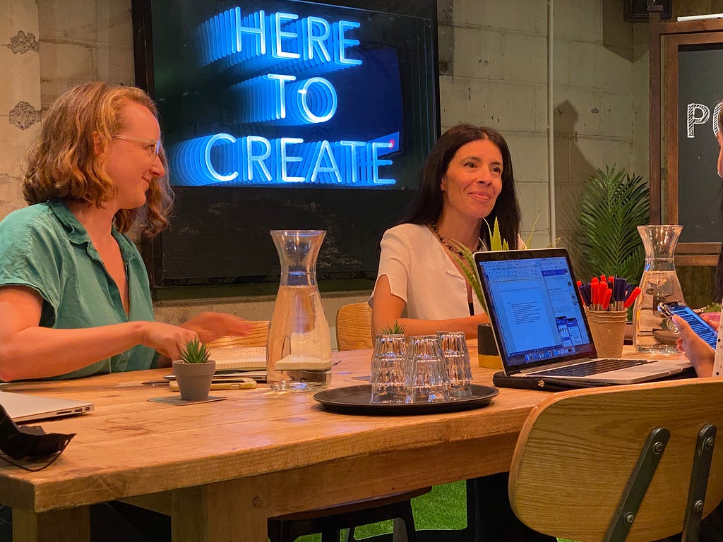 Megan and Rocio work at a table with a computer in front of them, with a neon sign saying “Here to Create” behind them.