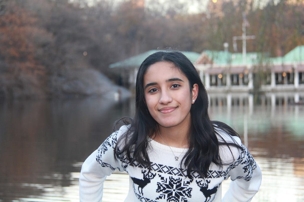 A young woman with brown skin in a white and blue reindeer sweater stands in front of a lake during fall.