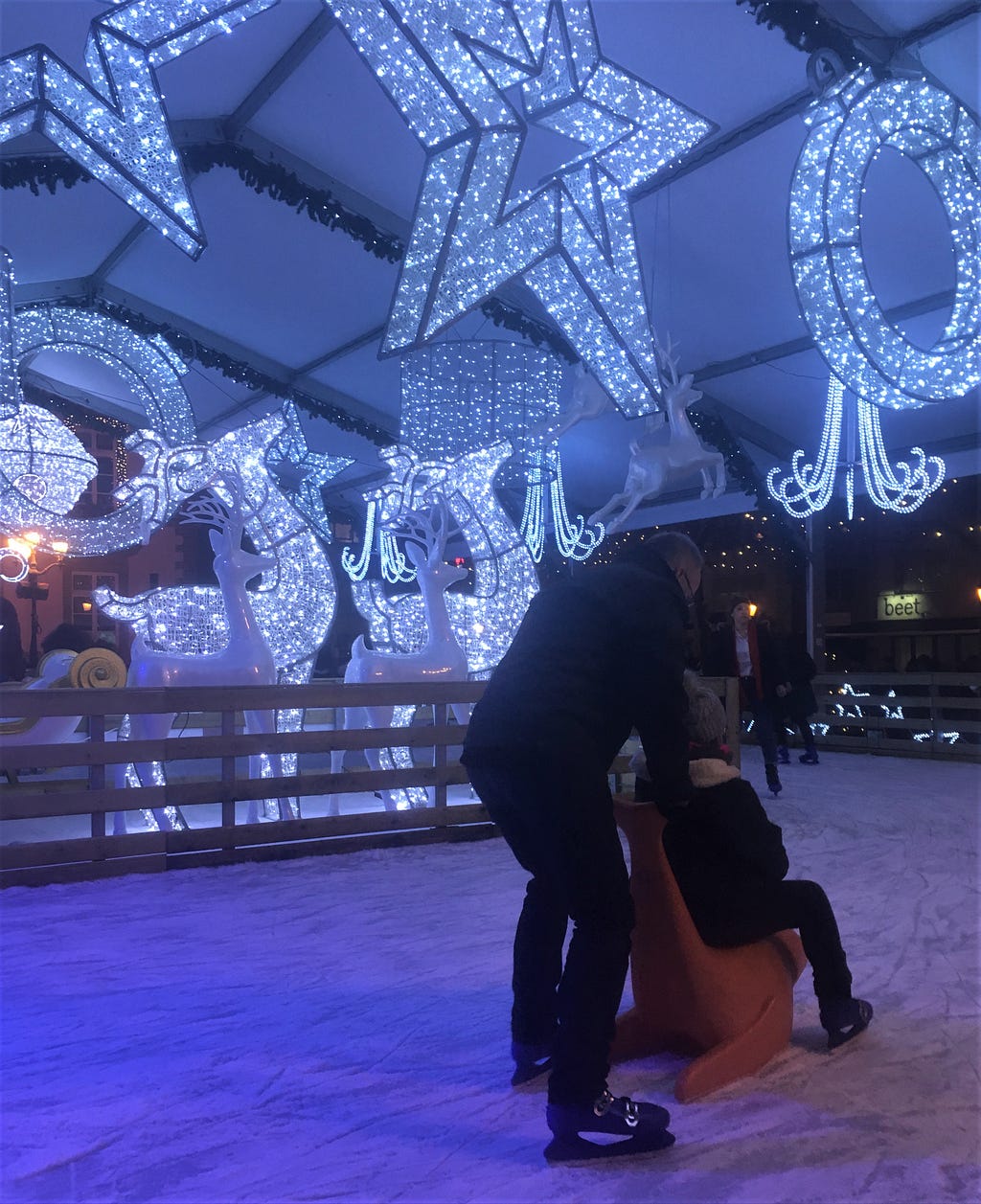 A person pushing another across an ice rink. Light structures of stars and deer in the background.