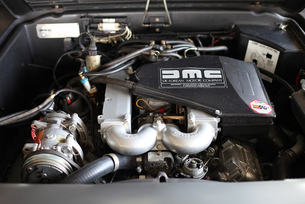 La imagen muestra un primer plano del compartimento del motor de un DeLorean, con el nombre “DMC DELOREAN MOTOR COMPANY” en la cubierta. Se pueden ver varios componentes alrededor del motor, incluyendo tuberías y cables.