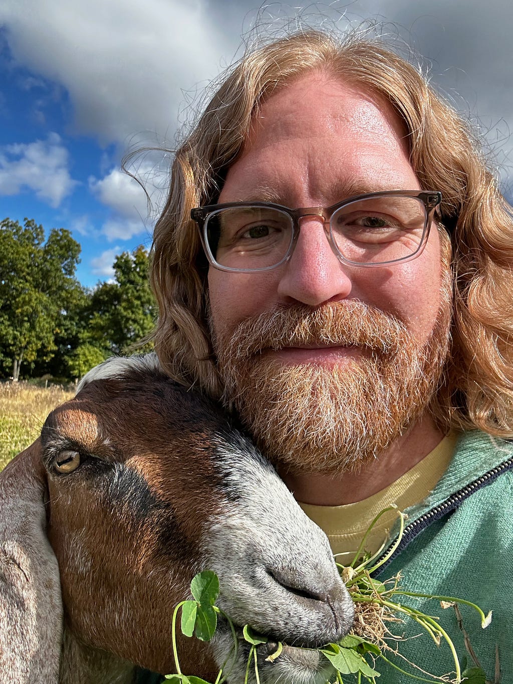 Selfie of a man with shoulder-length red hair and a beard, with a smattering of white hairs throughout. He is wearing brown glasses and looking at the camera. Below the man is a Nubian goat, with reddish fur and a white muzzle. The goat has a mouth full of clover. Behind them are a blue sky, trees, and a field.