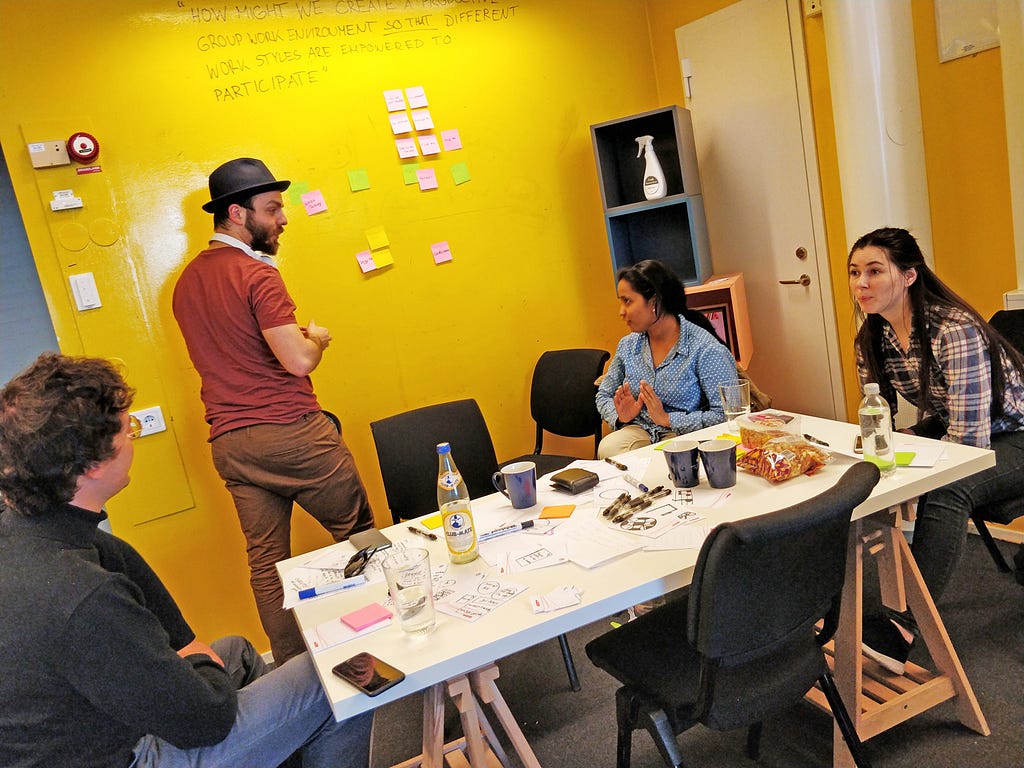 Four students in conversation, one is standing at the wall, placing Post-It notes in a grid.