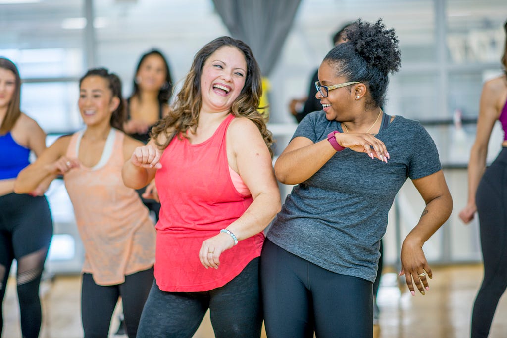 A group of people move joyfully in a fitness class. Everyone has big smiles and is dressed comfortably.