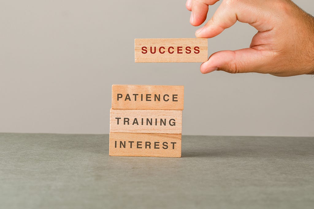 A hand stacks small wood blocks on top of each other. From bottom to top: “Interest,” “Training,” “Patience,” “Success.”