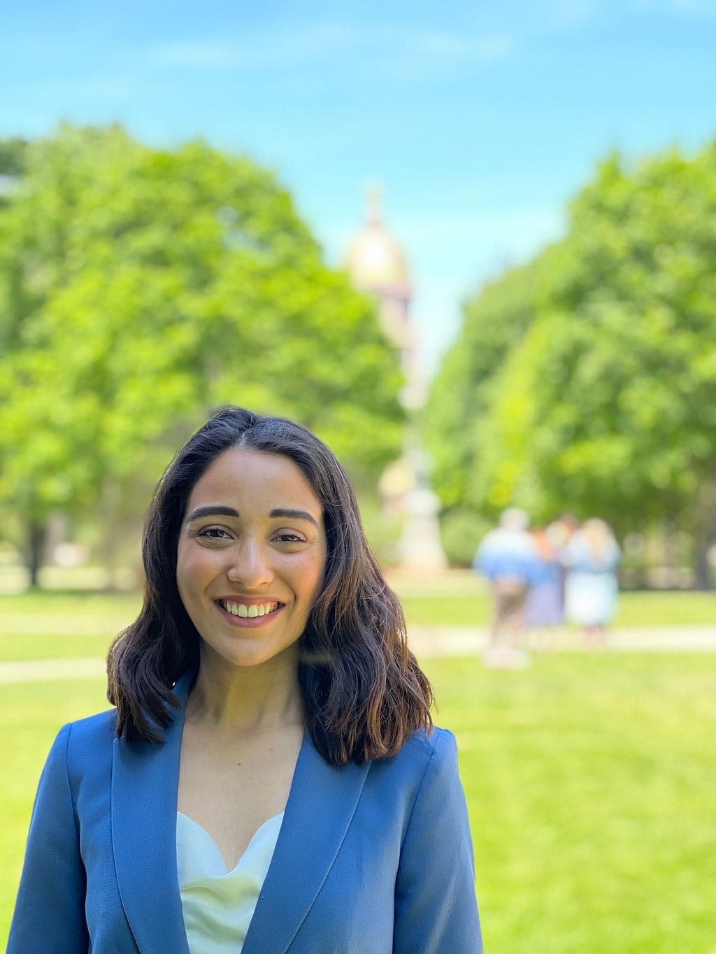 Marla standing in the greenery of her campus wearing a grey suit.