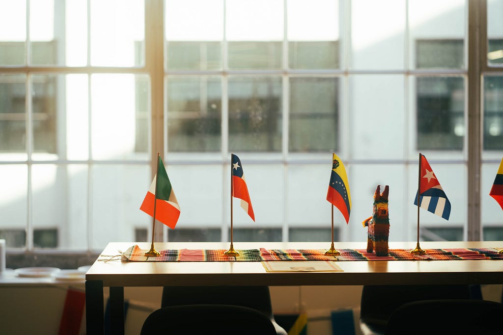 Small flags representing Spanish-speaking countries sit on top of a long, narrow table. In the background, light filters through big warehouse windows.
