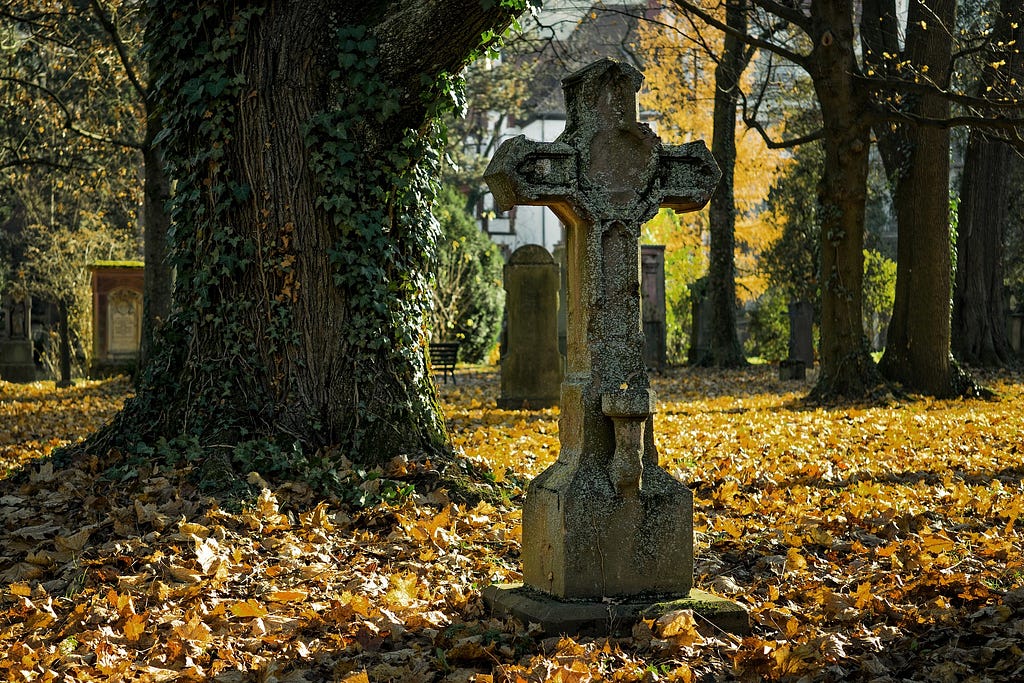 A cross-shaped tomb stone next to an old tree in an autumnal graveyard.