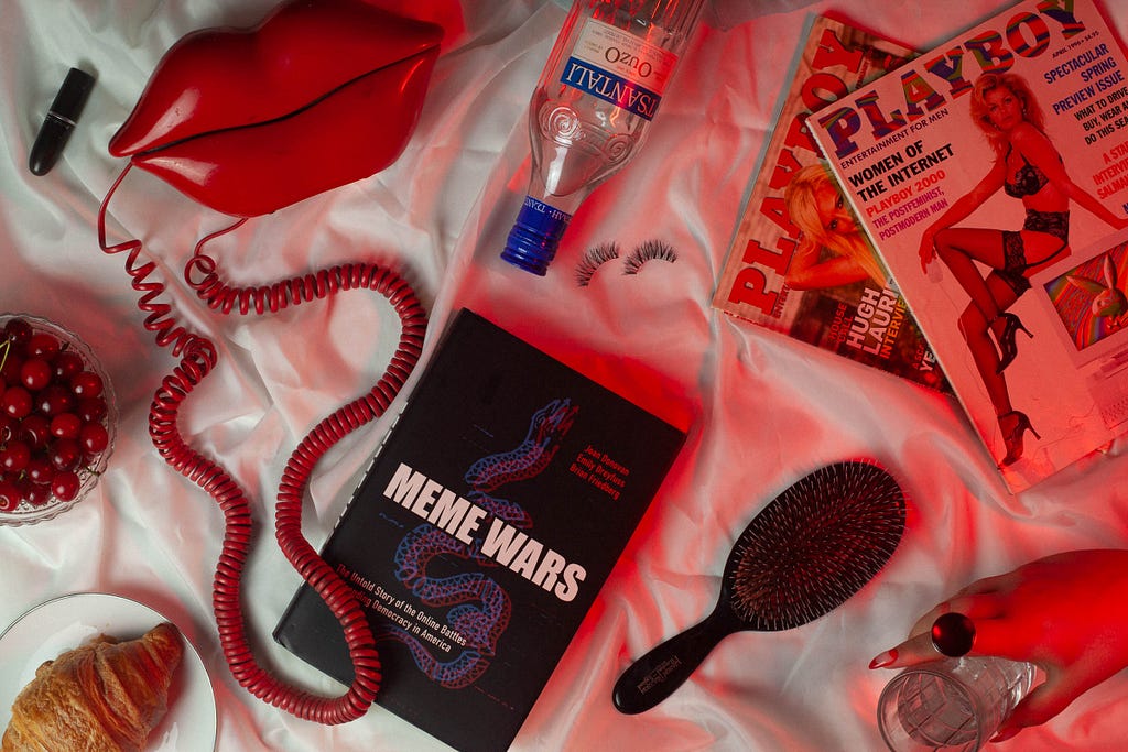 An eclectic scene on a white satin background includes a red lips phone with a coiled cord, a bottle of Iskilde water, a black hairbrush, false eyelashes, two Playboy magazines, a book titled “Meme Wars,” a bowl of cherries, a croissant on a plate, a black lipstick, and a hand holding a glass. Red lighting casts a dramatic hue over the setup.