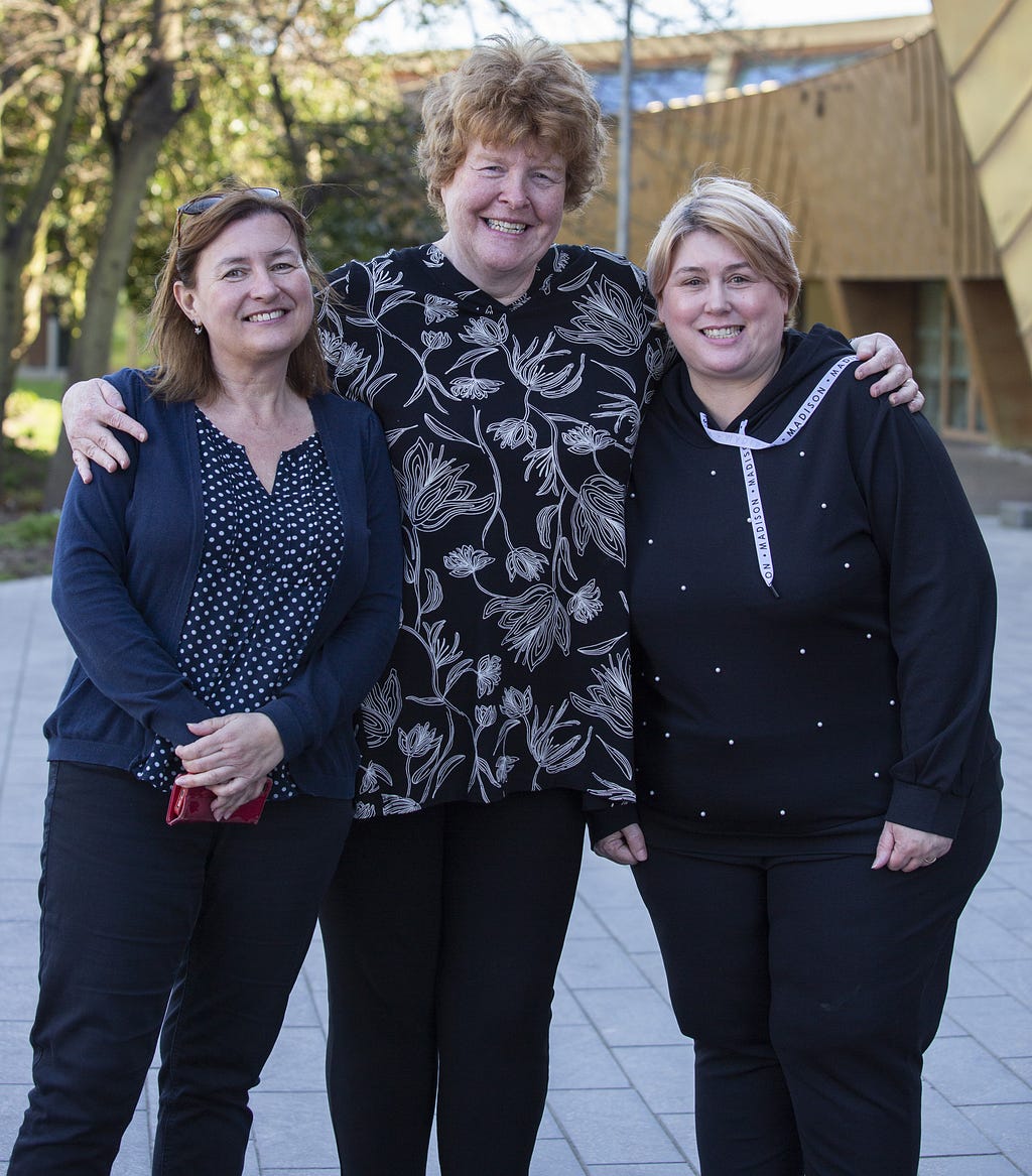 Three women stand smiling outside of a building