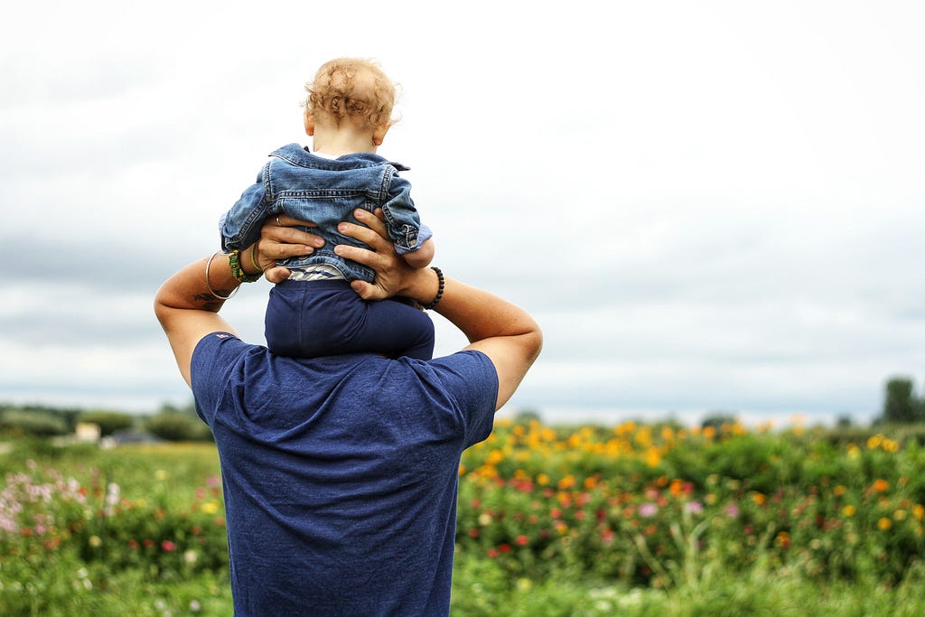 Pai carrega filha bebê sentada em seus ombros no meio de um campo de flores. Esta foto ilustra um texto sobre como obter residência permanente na Estônia.