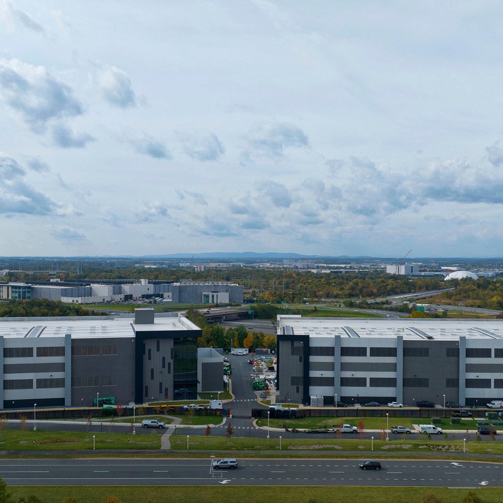 Aerial view of a large data center complex with modern buildings, parking lots, and surrounding greenery. The sky is overcast, and the horizon shows more industrial buildings and infrastructure.