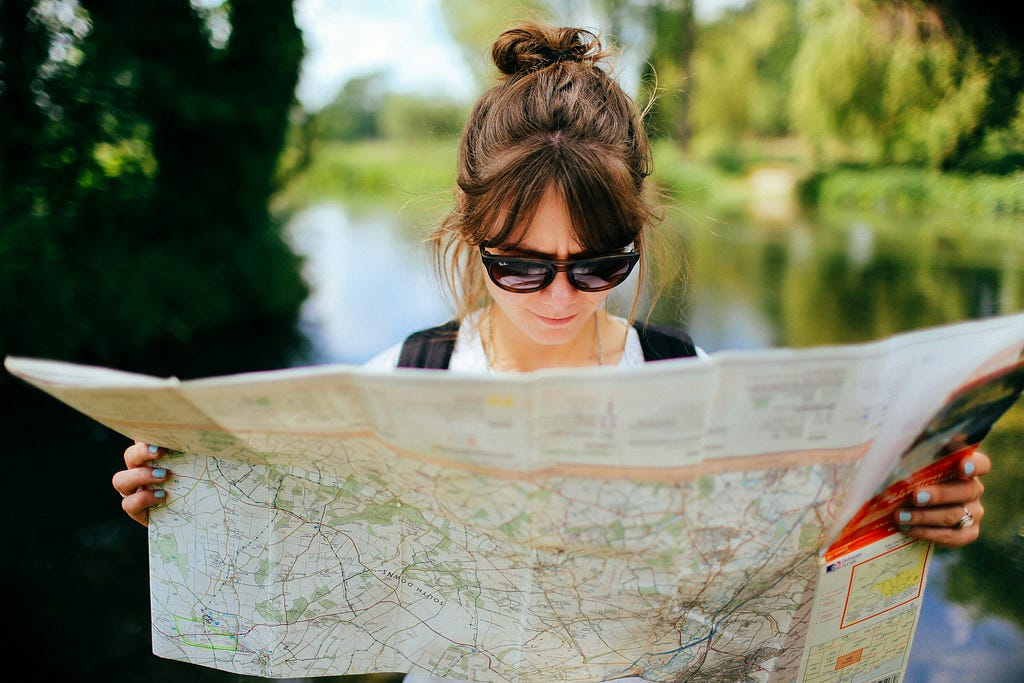 Woman with hair in a bun and sunglasses reading an unfolded paper map outdoors, with a blurred green park and water visible in the background