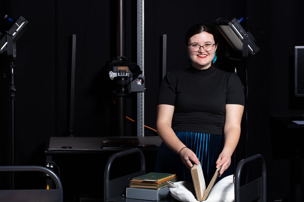 Image is of a person with glasses holding a book on a trolley in front of two large photography studio lights, a table and a mounted camera.