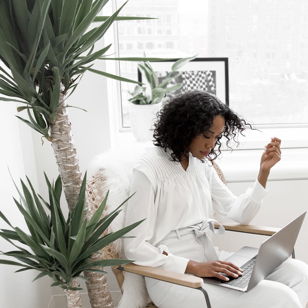 A woman dressed in white blouse and grey pants sitting on a chair looking at an open laptop. Large green plant to her left side and a large window behind her.