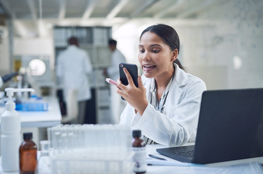A woman in a lab coat applies lip gloss while staring at her phone. She is at her desk in a laboratory.