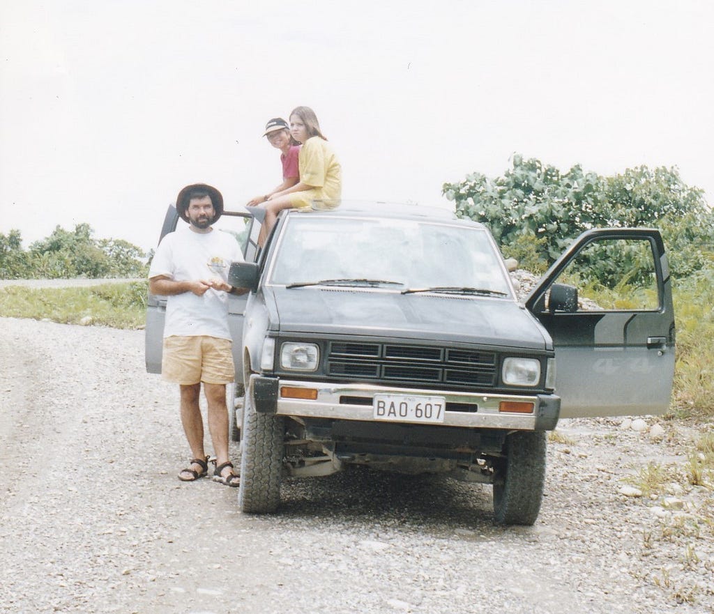 The author’s father in a tilly hat, shorts and sandals stands to the left of a Toyota 4x4 pickup. Two kids sit on the roof of the truck, one grinning, one scowling.