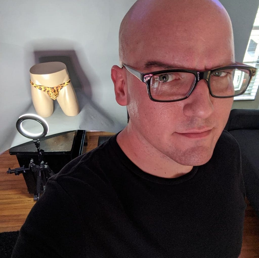 A white, bald man in glasses and a black t-shirt stands in front of a mannequin pelvis, illuminated by a ring light, wearing a leopard print thong.