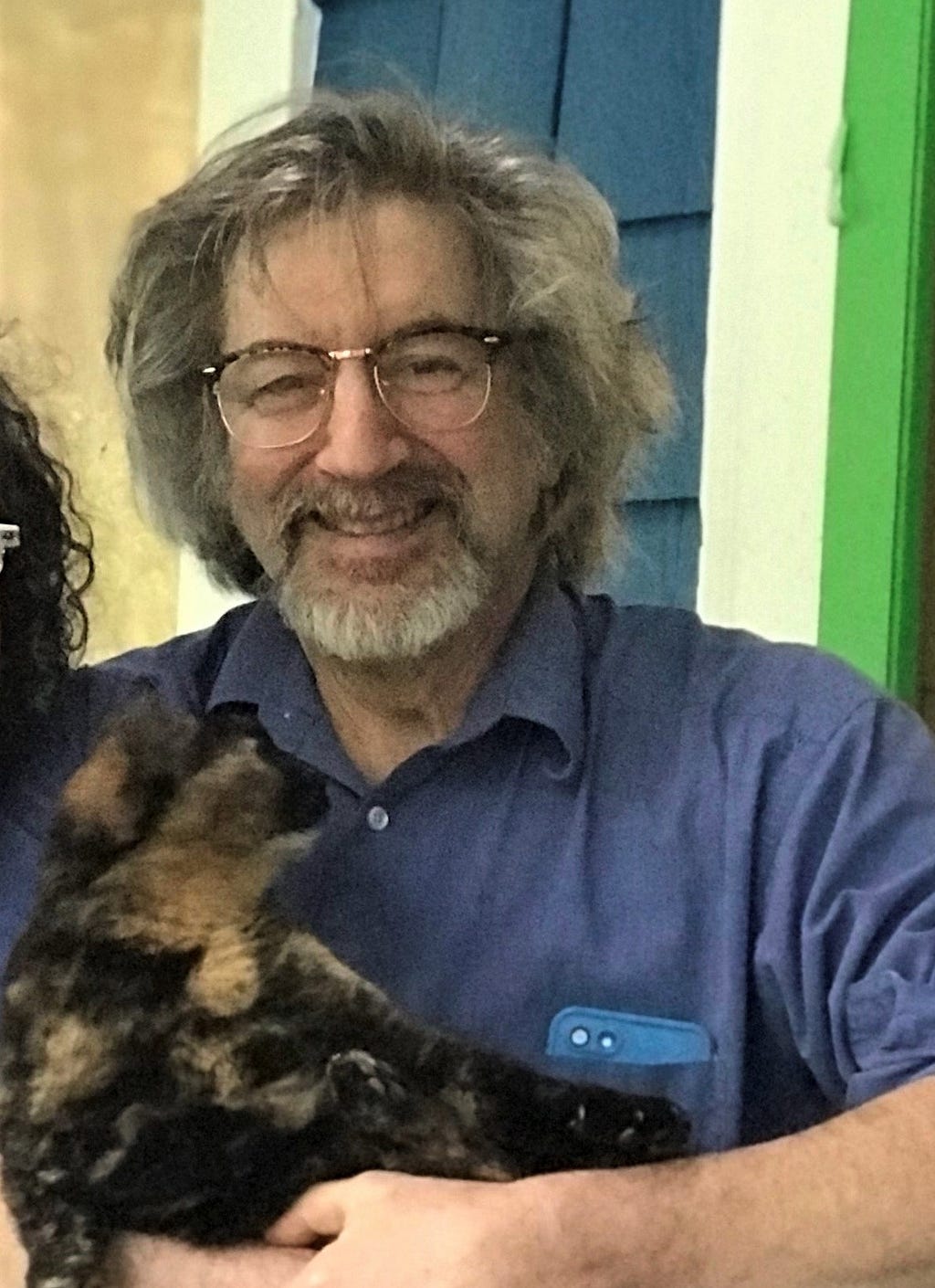 A man with medium-length gray hair, gray beard and mustache, glasses, and blue shirt smiles while holding a black-and-orange cat.