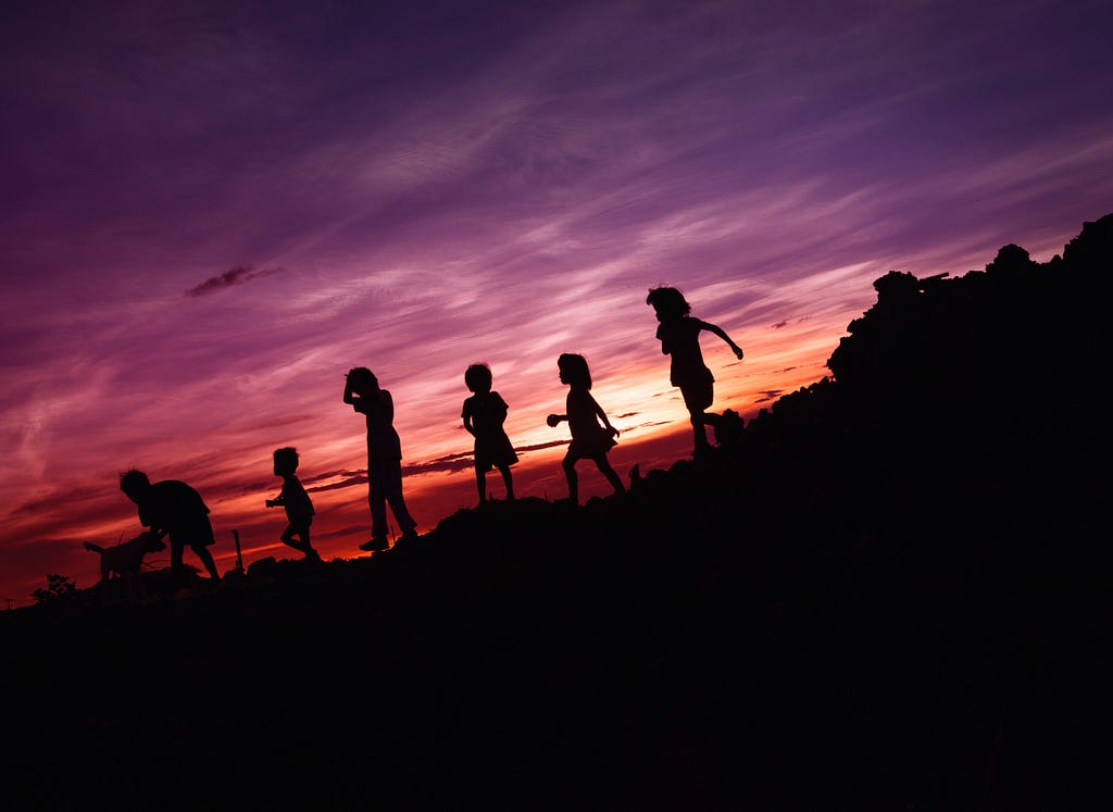 Children running along a hillside are silhouetted by a sunset
