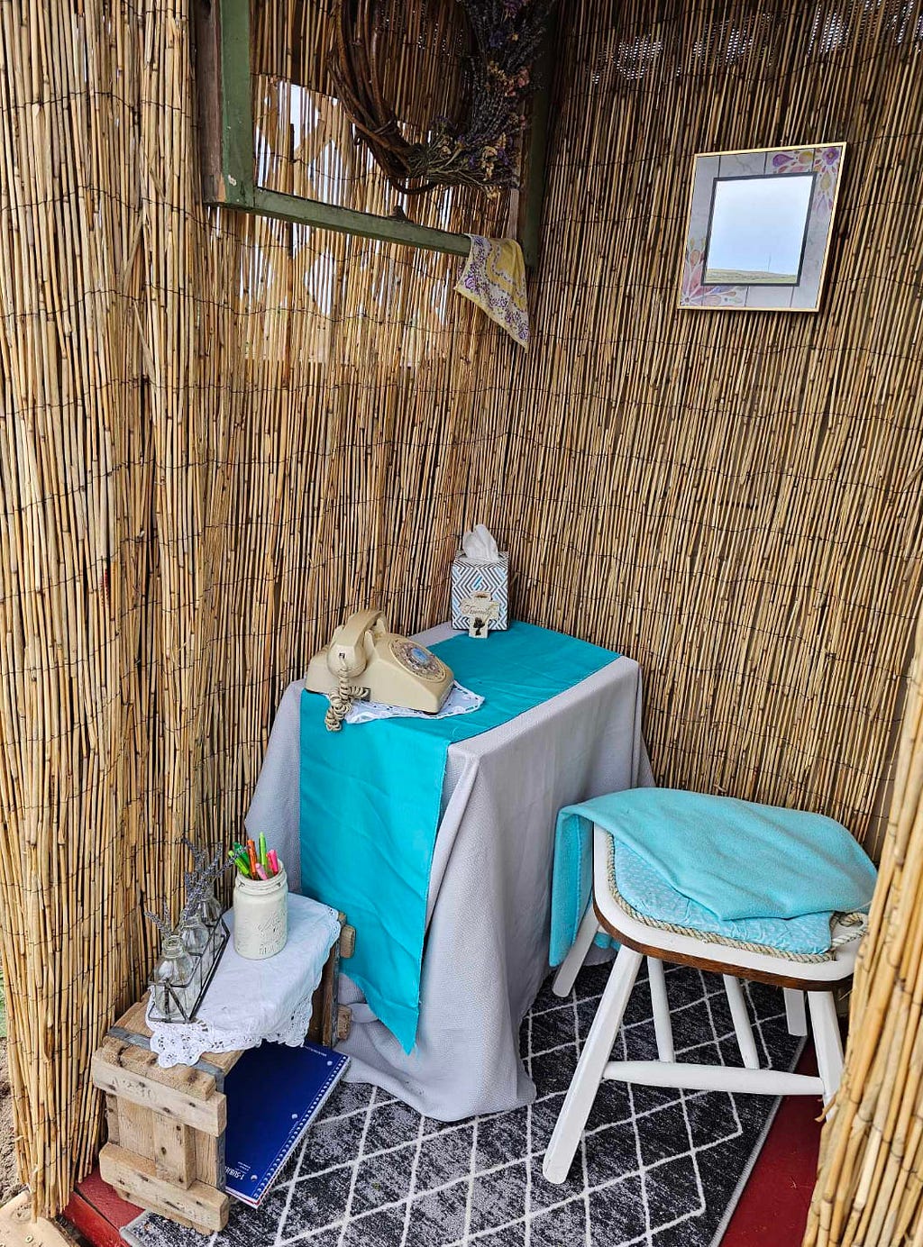 Booth with a white chair and blue cloth on it in front of a table covered with a white cloth and blue liner and an ivory rotary phone and box of tissue and a small table to the left containing a jar of pens, smaller glass jars and notebooks underneath.