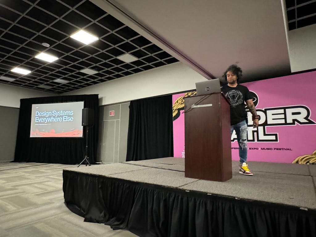 Dan Mall giving a presentation on Design Systems on a small stage in front of a large banner with pink and white lettering.