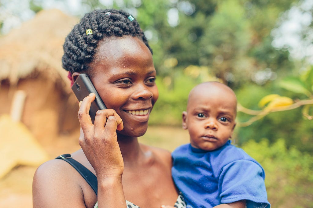 A smiling woman holds a mobile phone to her ear while also holding a small child in her other arm.