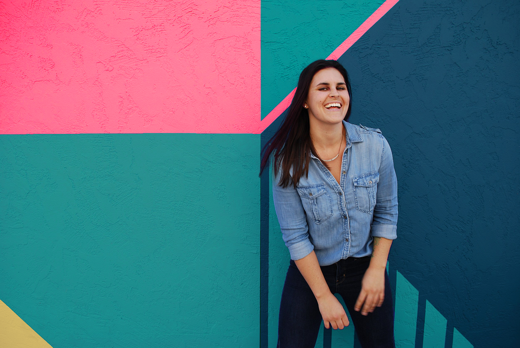 A young woman in a denim shirt and dark pants in motion and smiling in front of a colorfully painted wall
