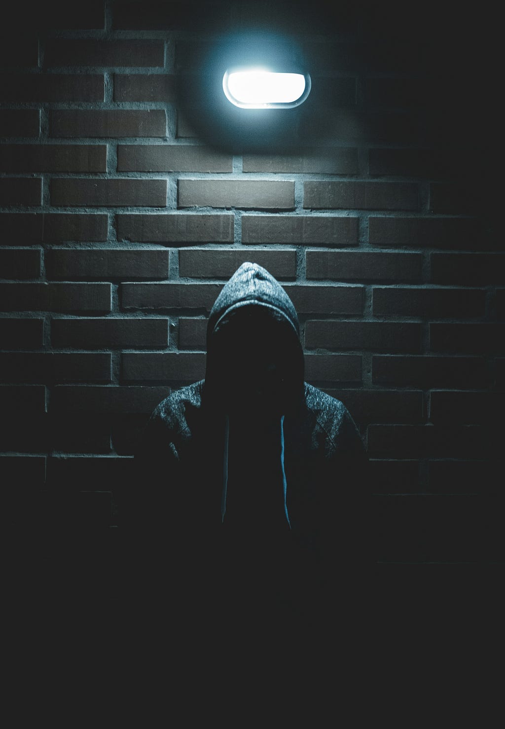 A figure in a hoody standing in front of a brick wall. They are backlit so that they face is in shadow.