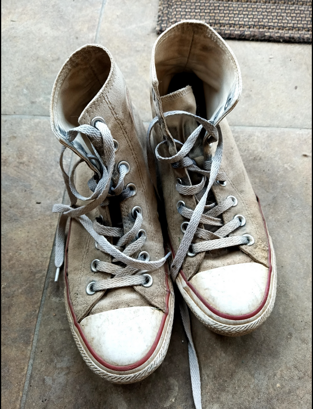 A pair of sneakers, Converse high tops, dirty and worn, rest on a tile floor.