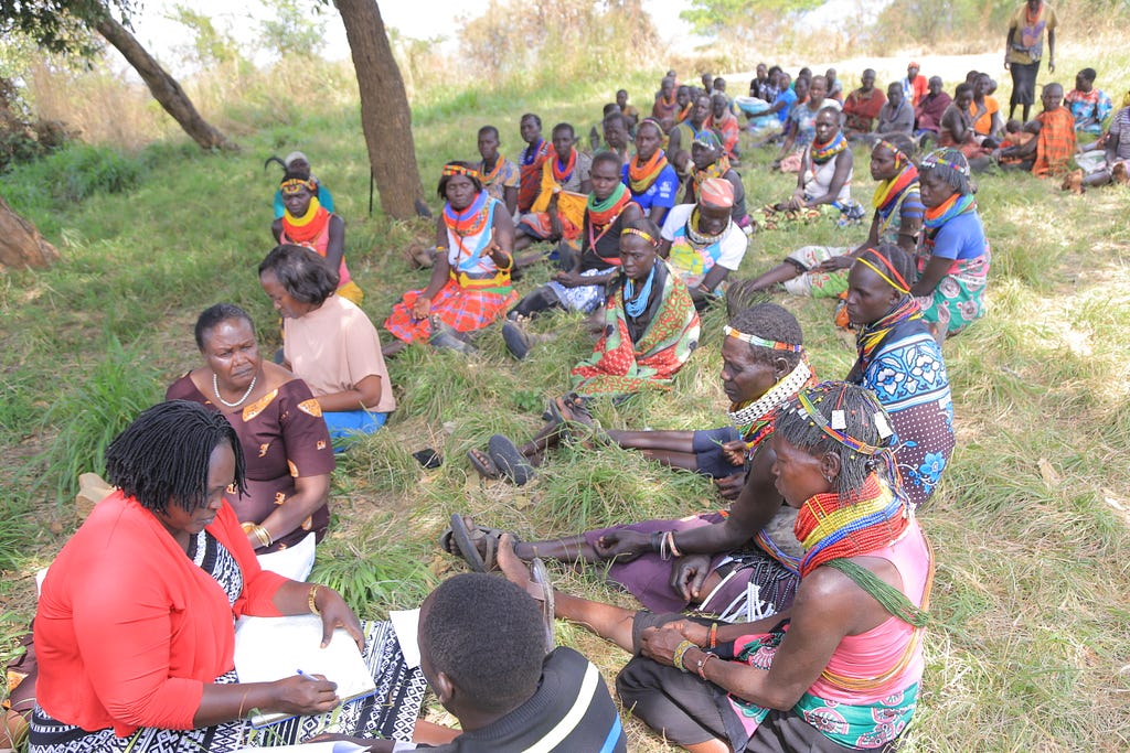 A researcher sits outside on the ground beneath trees with papers on her lap with a line of Indigenous people sitting on the ground stretched out before her.