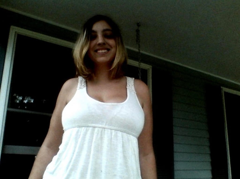 A young white woman wearing a white tank top smiles into the camera. The background is dark and her eyes are not clearly visible, but there are shutters and the siding of a house in the background.