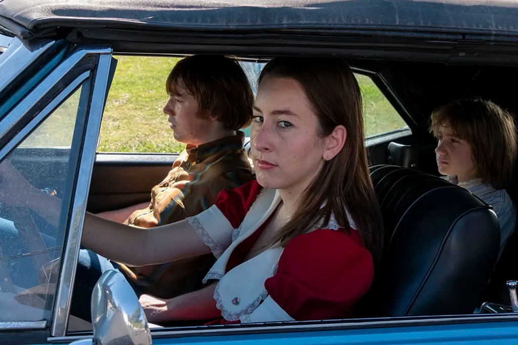 Alana (Alana Haim), Gary (Cooper Hoffman), and his younger brother are sat in a convertable car. The two boys look ahead as Alana, who is driving, breaks the fourth wall by staring into the camera.