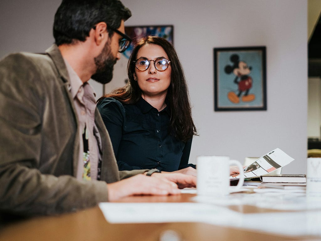 Two people sit at a meeting table reviewing documents in an office setting, with a framed picture of Mickey Mouse hanging on the wall in the background and coffee mugs placed on the table.
