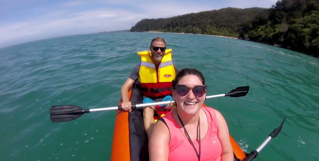 A smiling woman sat in front of a man wearing sunglasses and a life jacket. Both are smiling. They are in an inflatable kayak, floating on blue water.