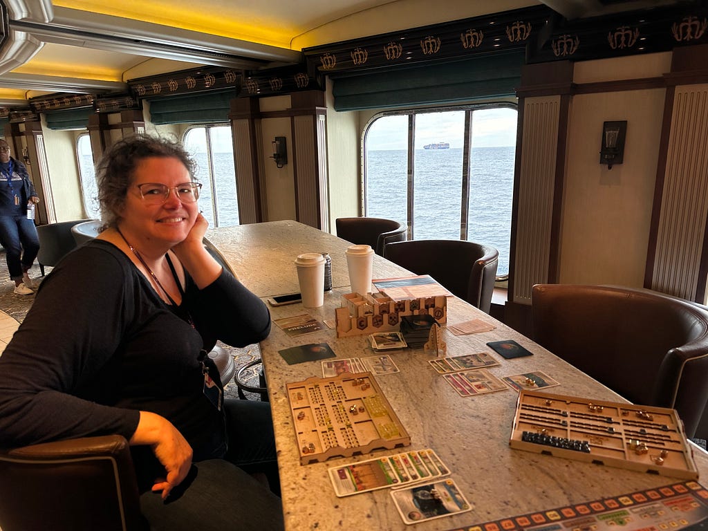 A smiling middle aged woman with glasses and frizzy hair pulled back in a pony tail sits in front of a very long table with a bunch of board game components on it, with a large picture window in the background that has a view of the ocean and another ship in it.