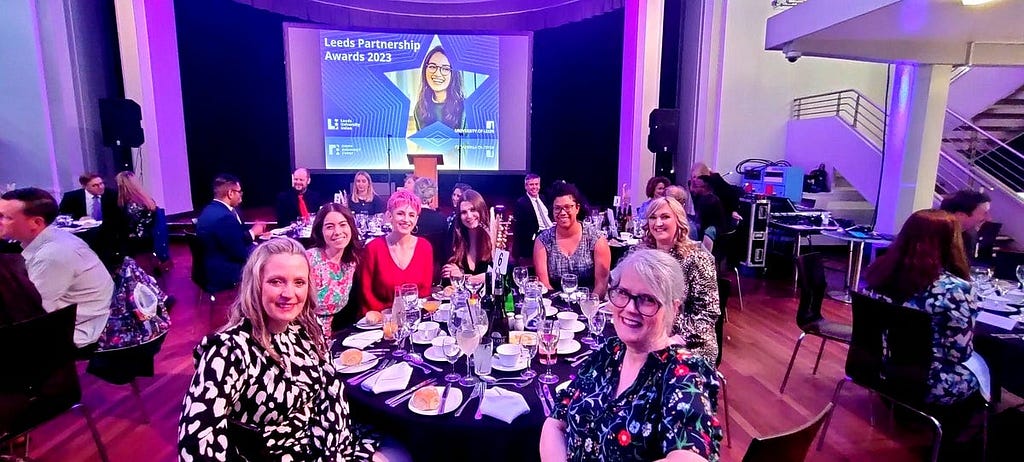 An image of Emma Taylor, Laura Conroy, Lauren Huxley, Zofia Wilk, Bryony Brown, Kim Crossley and Jo Huett sat at a circular table in front of a stage where there is a screen displaying an image of a student and text which reads ‘Leeds Partnership Awards 2023’.