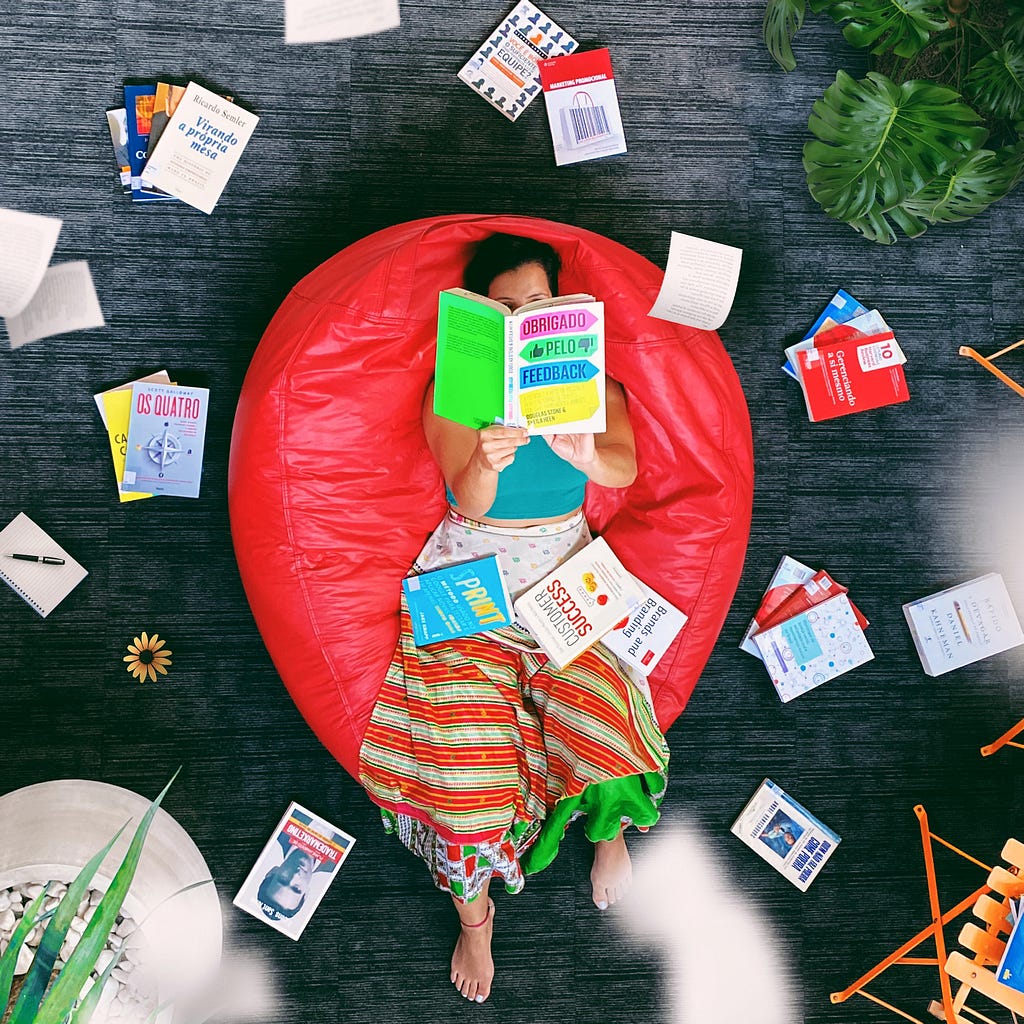 Puff vermelho com uma mulher branca com cabelo preto lendo um livro colorido e ao redor diversos livros espalhados.