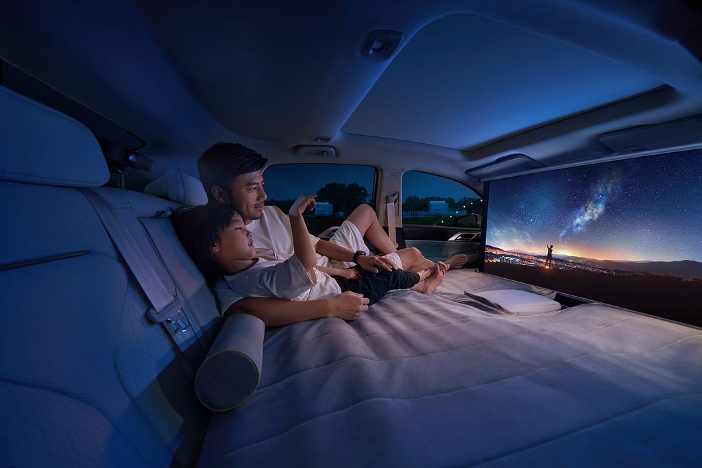 A father and child relaxing in the back of a car, transformed into a bed mode, watching a scenic night sky on a large screen.