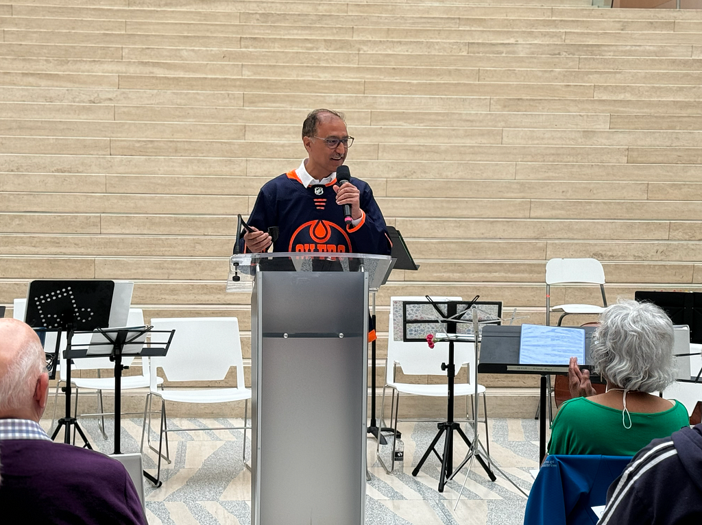 Mayor Sohi is at a lectern in the atrium of City Hall. He is wearing an Oilers Jersey as he addresses Edmonton Seniors seated infront of him.