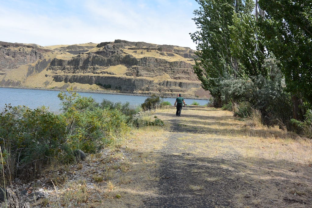 Photo of a person walking on path by the side of a lake, with trees and cliffs in the background