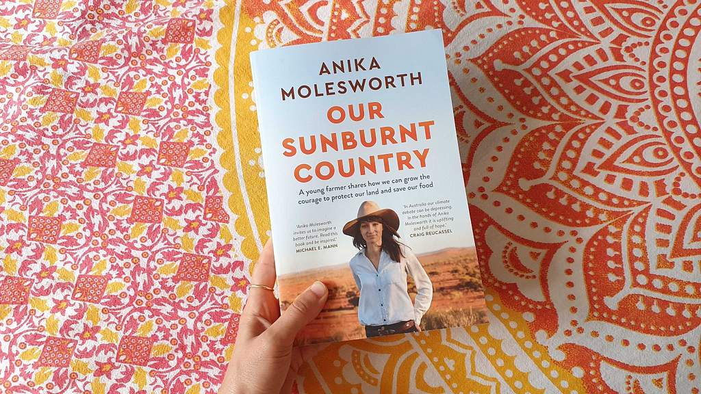 the review writer’s hand holding a copy of Our Sunburnt Country in front of a beautifully patterned orange and fabric gold background