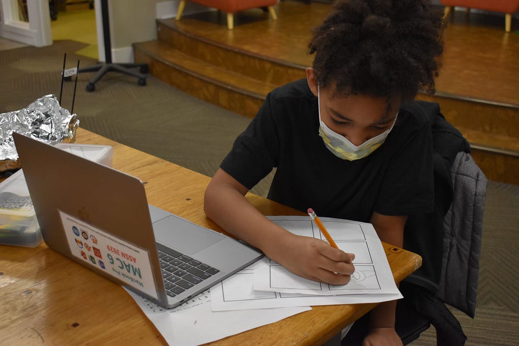 A young student sits at a computer and writes on a piece of paper. They are participating in Spark Central’s Level Up program.