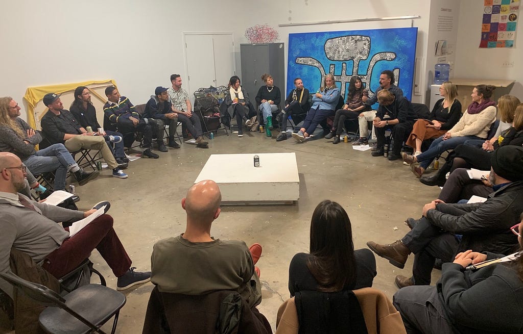 A group of 30 people sit in a circle. There is a low white square table in the center of the room and a large artwork with a black and white drawing against a bright blue background.