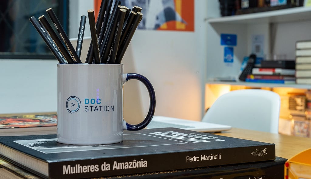 Imagem de um copo com o nome da DocStation escrito e com o seu logo. O copo está sobre uma pilha de livros, como o Mulheres da Amazônia, de Pedro Martinelli.